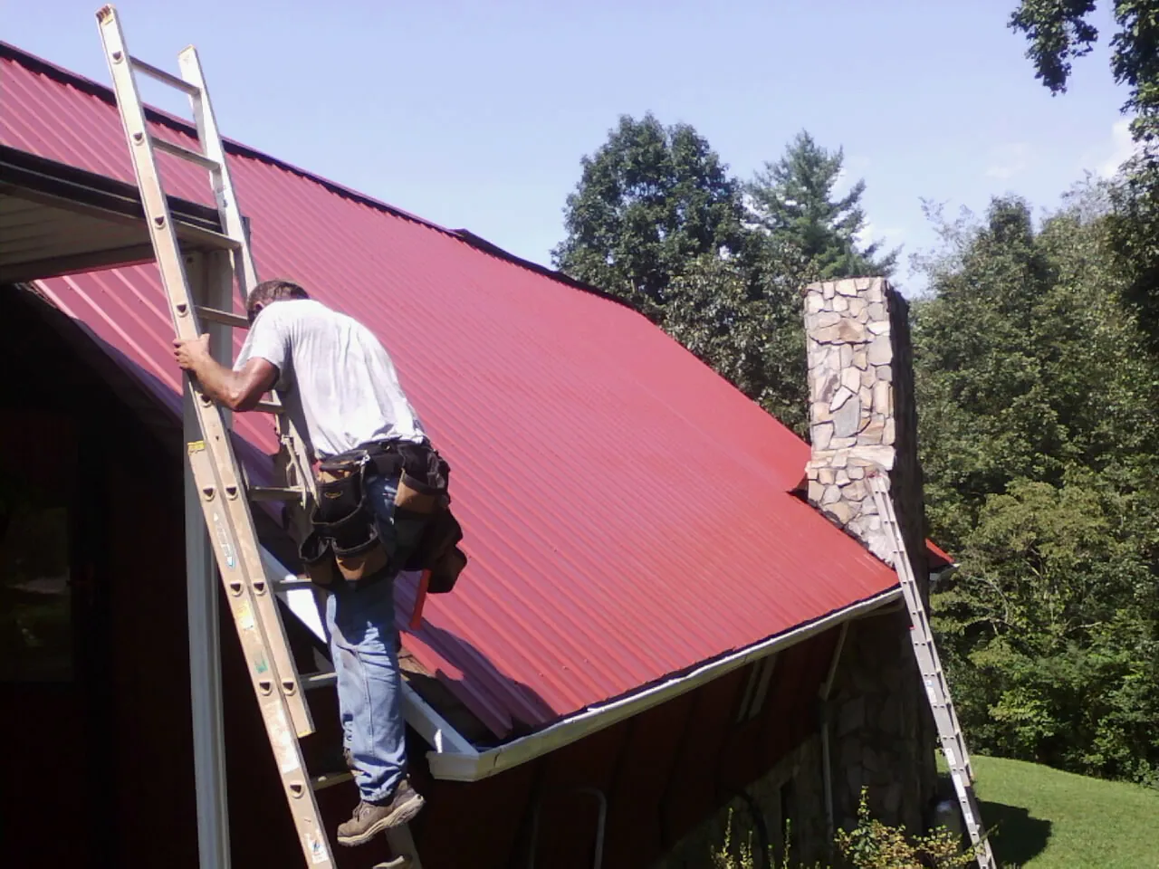 Roofer on a ladder beside a red metal roof and stone chimney — Soffit & Fascia Repair by Roofing Brothers in Greeley